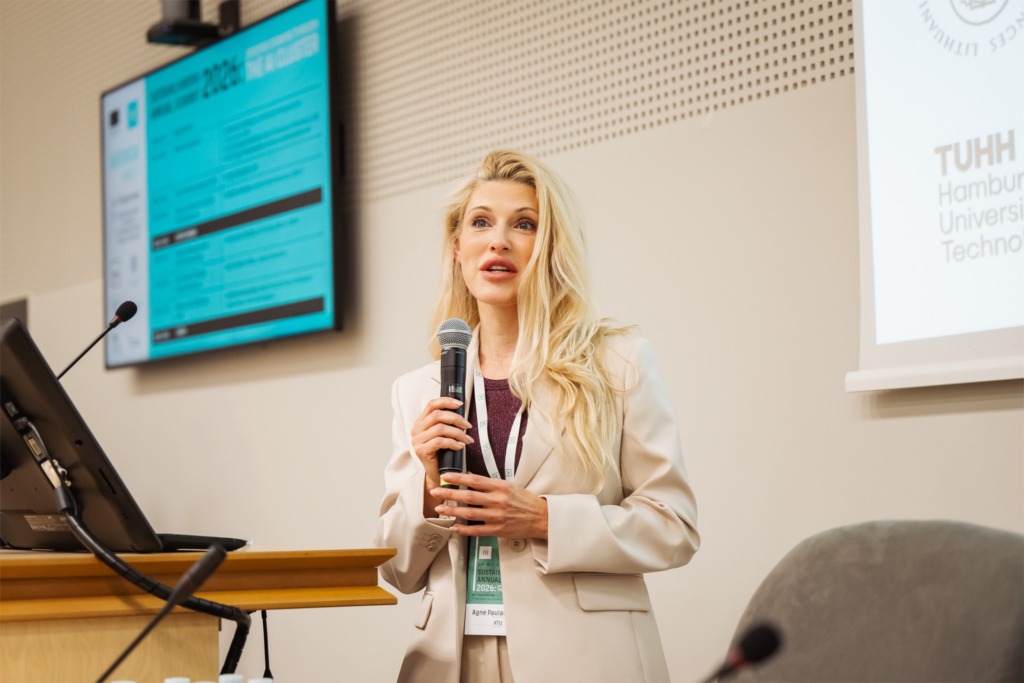 Prane&scaron;ėja su mikrofonu skaito prane&scaron;imą SustAInLivWork Annual Summit 2026 renginyje. Už jos ekrane matomas Hamburgo technologijos universiteto pavadinimas. Renginyje susitinka skirtingų sektorių dirbtinio intelekto ekspertai.