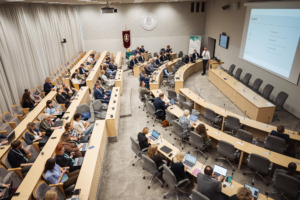 SustAInLivWork Annual Summit 2026 dalyviai klausosi pranešimo apie duomenų infrastruktūrą ir dirbtinio intelekto sprendimus. Auditorijoje susėdę renginio dalyviai stebi salės priekyje vykstantį pristatymą.
